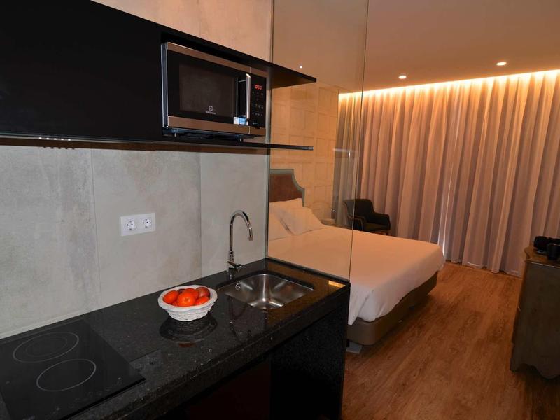 Komfortowy pokój hotelowy z aneksem kuchennym, podwójnym łóżkiem i ciepłym oświetleniem przy zasłonach.