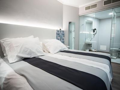 Habitación de hotel moderna con cama grande, paredes blancas y baño moderno con mampara de vidrio.