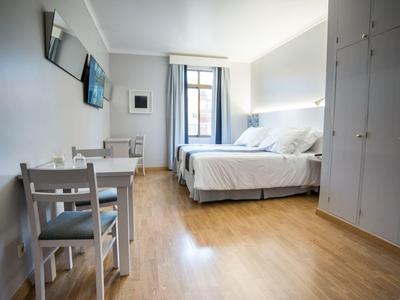 Habitación de hotel luminosa con suelo de madera, cama doble, armario, mesa y dos sillas junto a ventana.