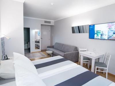 Habitación de hotel moderna con cama doble, sofá gris y televisor de pantalla plana en espacio iluminado.