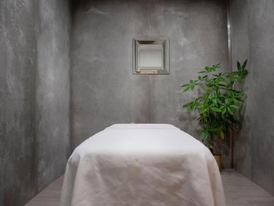 Jednoduchá spa místnost se šedými zdmi, masážním stolem a květinou v rohu.