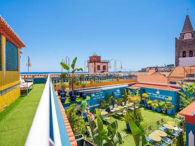 Terraza en la azotea con asientos, plantas y vista al mar y edificios históricos bajo cielo despejado.