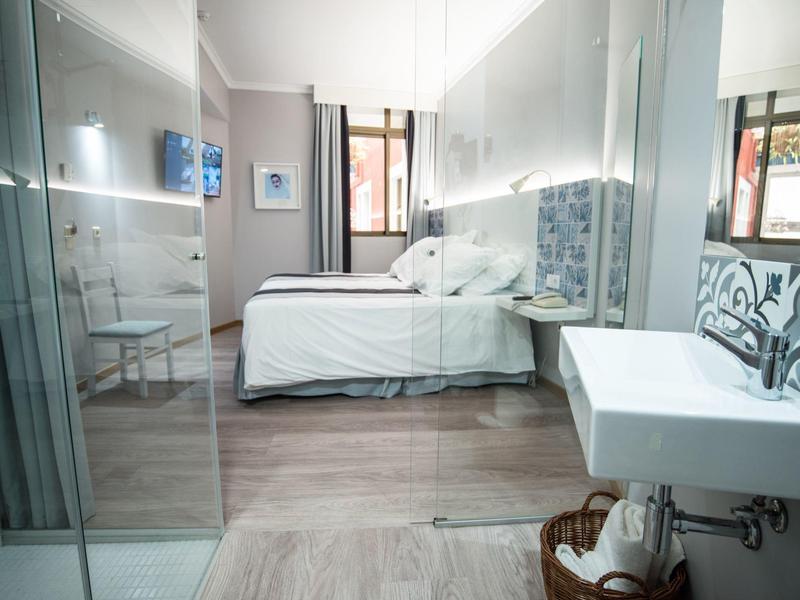 Habitación de hotel moderna con cama doble, ducha de vidrio y lavabo grande en diseño claro.