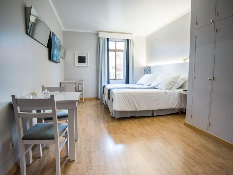 Habitación de hotel luminosa con suelo de madera, cama doble, armario, mesa y dos sillas junto a ventana.