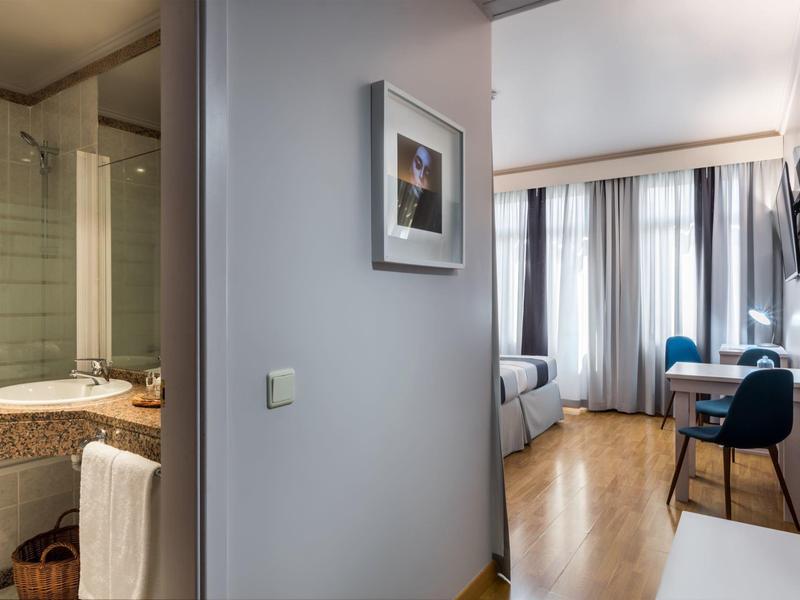 Pasillo de habitación de hotel moderna con vista al baño y área de estar con piso de madera y grandes ventanas.