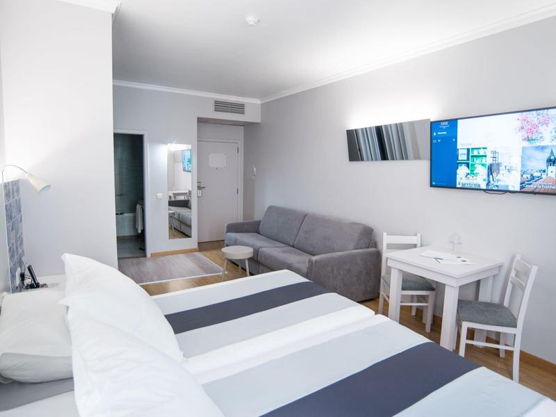 Habitación de hotel moderna con cama doble, sofá gris y televisor de pantalla plana en espacio iluminado.