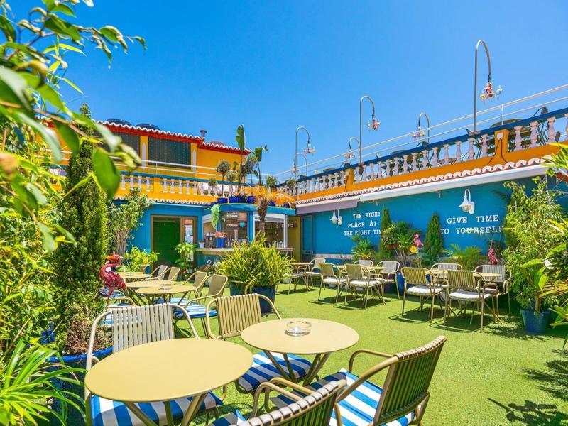 Colorida terraza de hotel con mesas, sillas y plantas verdes bajo el cielo azul.