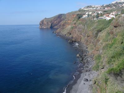 Küstenklippe mit blauem Meer, Felsenstrand und Häusern auf dem Hügel bei klarem Himmel.
