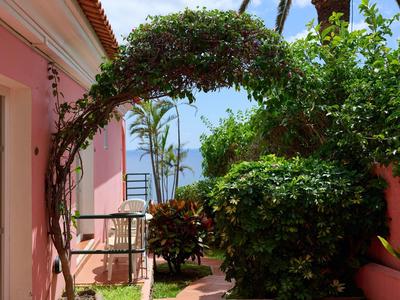 Grüner Pflanzenbogen über Gartenweg zwischen rosa Hauswänden bei sonnigem Himmel.