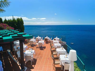 Terrasse mit weißen gedeckten Tischen und Stühlen an der Küste mit blauem Himmel und Meer im Hintergrund.