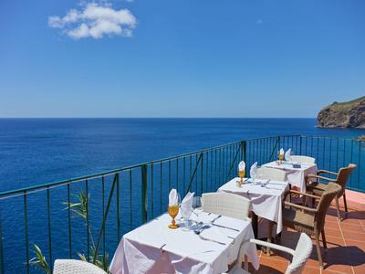 Terrasse mit weißen gedeckten Tischen und Stühlen am Meer unter blauem Himmel mit wenigen Wolken.