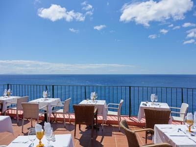 Terrazza soleggiata con tavoli e sedie con vista sul mare blu sotto un cielo limpido.