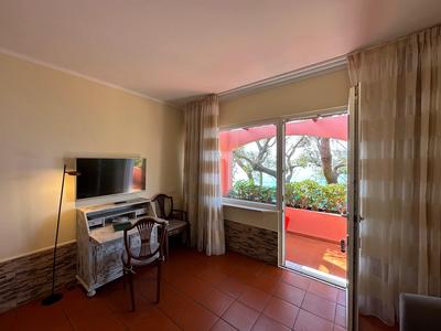 Helles Wohnzimmer mit Fernseher, Stuhl, Schreibtisch und Glas-Schiebetür zum Balkon mit rotem Sonnenschirm.