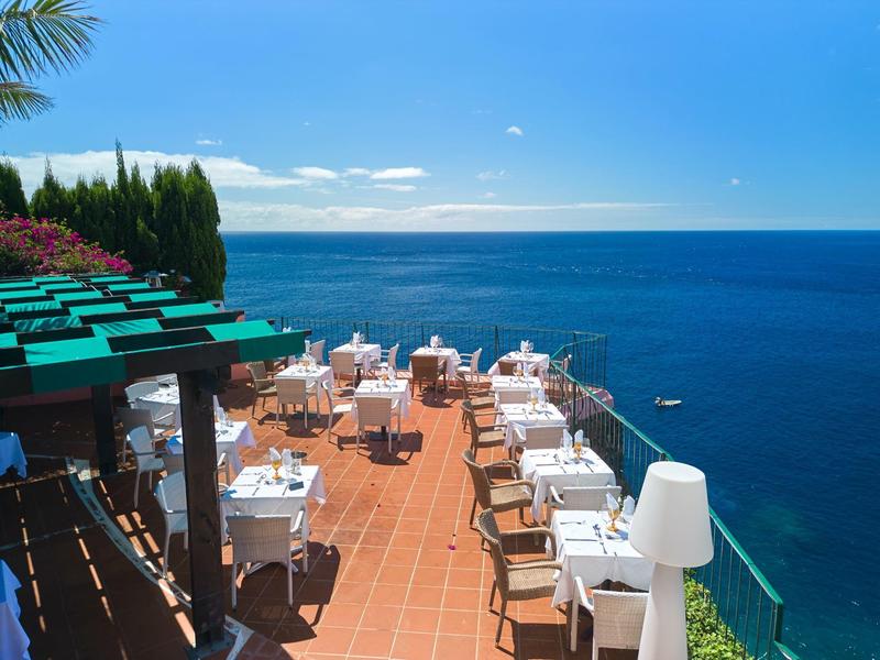 Terrasse mit weißen gedeckten Tischen und Stühlen an der Küste mit blauem Himmel und Meer im Hintergrund.