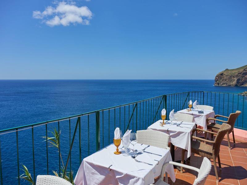 Terrasse mit weißen gedeckten Tischen und Stühlen am Meer unter blauem Himmel mit wenigen Wolken.