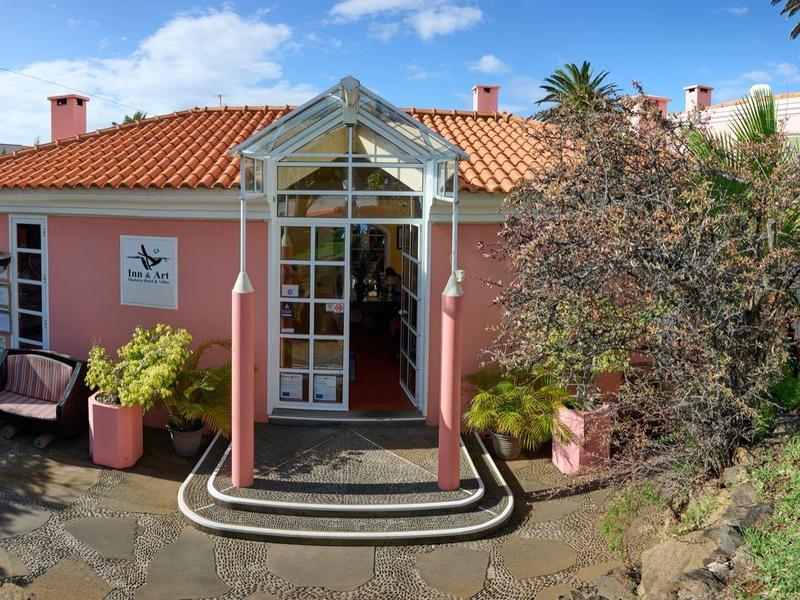 Ein rotes Haus mit Glas-Vordach, blauen Himmel, Pflanzen und Palmen im Garten.