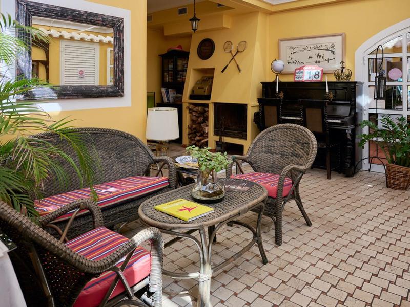 Gemütliche Terrasse mit Rattanmöbeln, gepolsterten Kissen, Pflanzen und gelber Wandgestaltung.