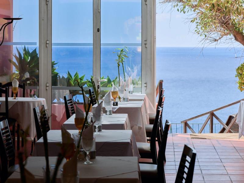 Restaurant mit weißen Tischdecken, Holzstühlen und Meerblick durch große Fenster und offene Tür.