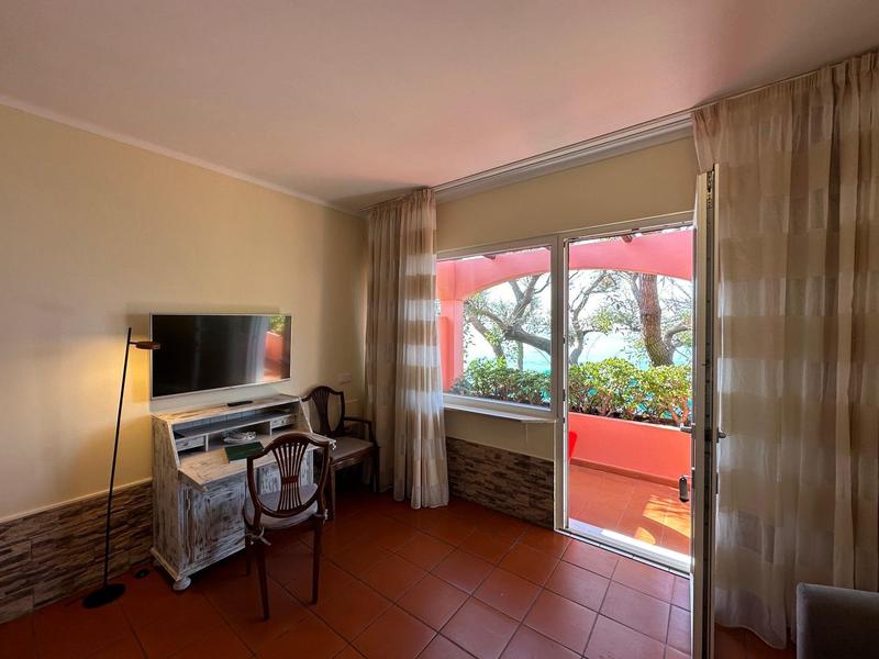 Helles Wohnzimmer mit Fernseher, Stuhl, Schreibtisch und Glas-Schiebetür zum Balkon mit rotem Sonnenschirm.