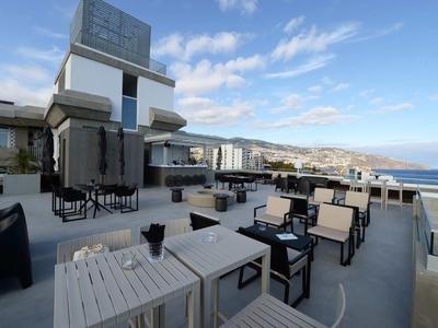 Buitenterras met moderne tafels en stoelen met uitzicht op zee en verre heuvels onder blauwe lucht.
