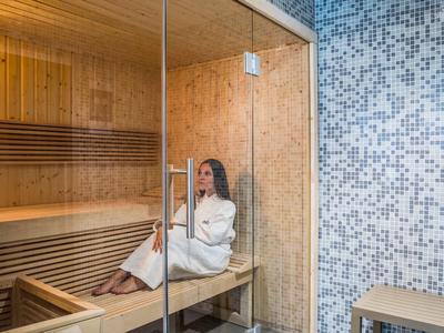 Vrouw in witte badjas ontspant in een houten sauna naast een betegelde muur.