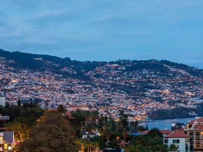 Stadslichten fonkelen op een kustheuvel bij schemering, met gebouwen en een baai zichtbaar.