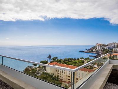 Blick von einer Hotelterrasse mit Glasgeländer auf das Meer und die Küstenstadt unter bewölktem Himmel.