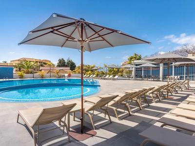 Poolbereich mit Sonnenliegen und Sonnenschirm auf einer Terrasse neben einem Hotel.