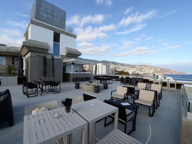 Buitenterras met moderne tafels en stoelen met uitzicht op zee en verre heuvels onder blauwe lucht.
