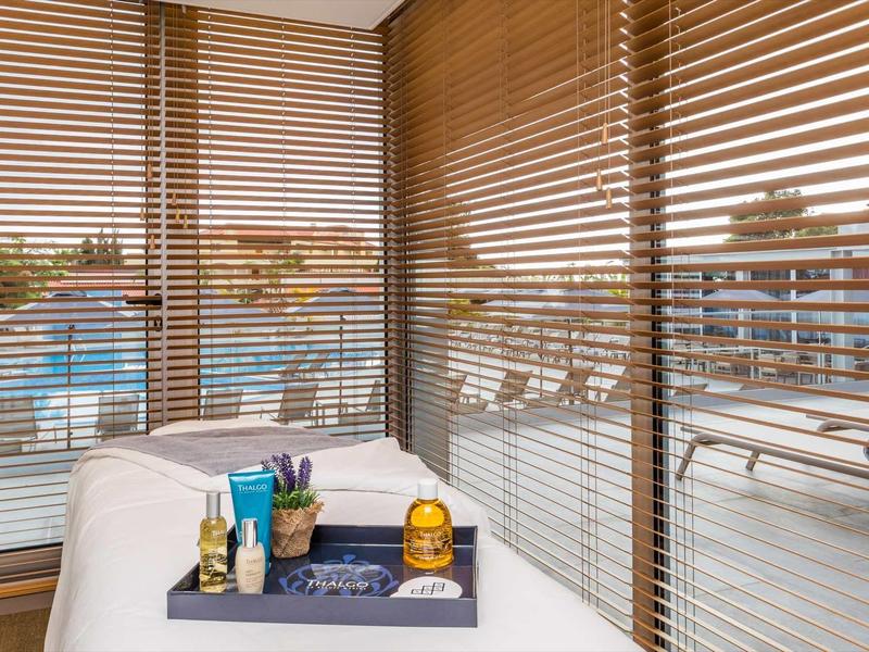 Hotelbalkon met houten jaloezieën, witte ligstoel en dienblad met toiletartikelen en kaarsen.