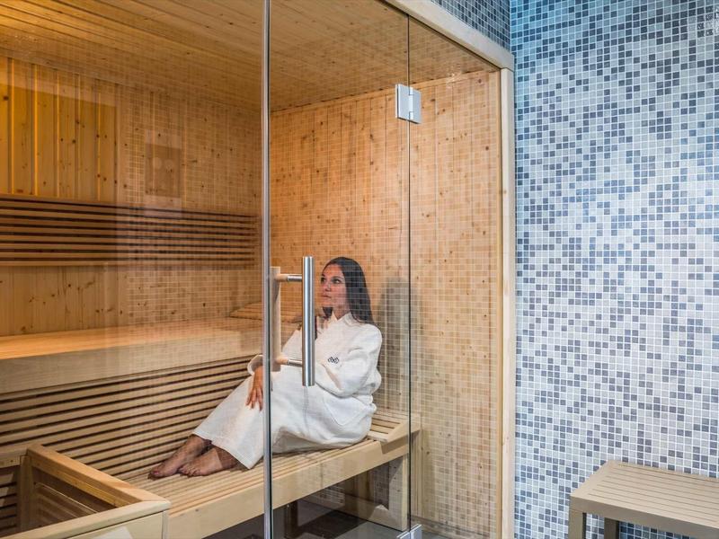 Vrouw in witte badjas ontspant in een houten sauna naast een betegelde muur.