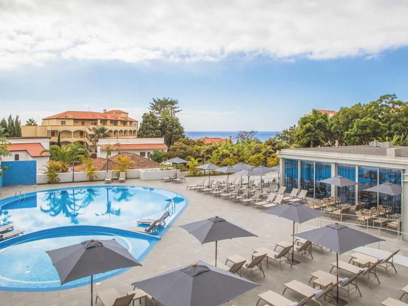 Buitenzwembad met parasols, ligstoelen en gebouwen onder een deels bewolkte lucht.