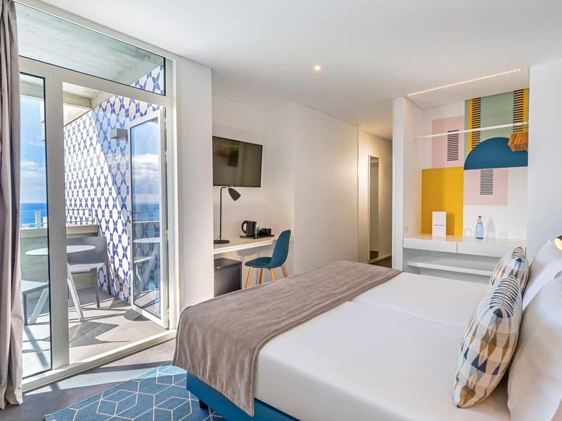 Lichte hotelkamer met tweepersoonsbed, open deur naar balkon en kleurrijke muurschildering.