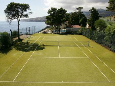 Tennis court with green surface and sea view, surrounded by trees and hills.