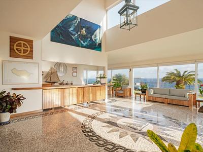 Jasna lobby hotelowa z recepcją i miejscem do siedzenia z widokiem na morze i roślinami.