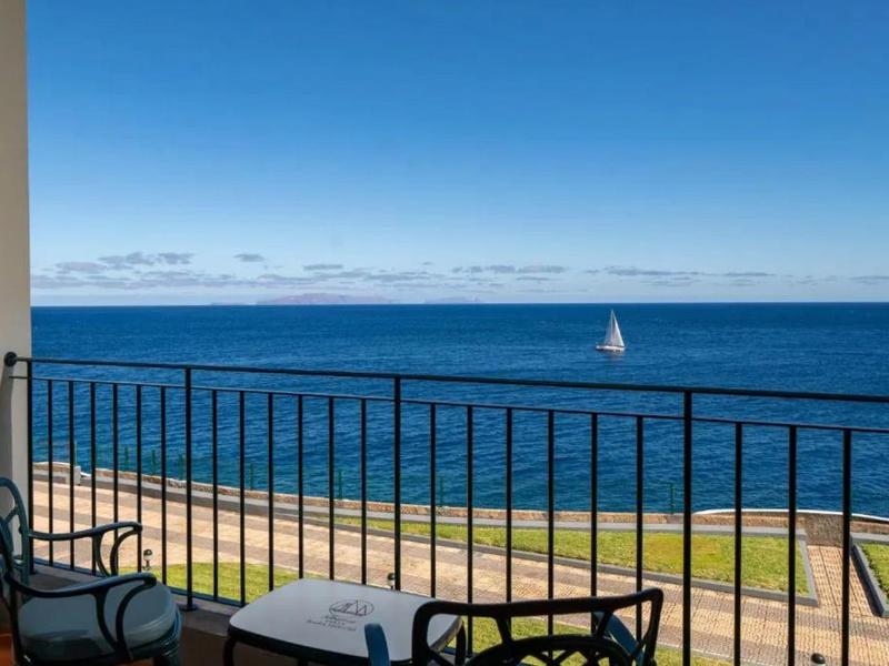 Balkon z stołem i krzesłami z widokiem na morze i żaglówkę przy czystym niebie.