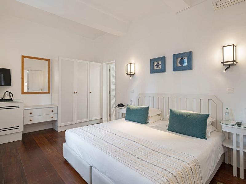 Prosto umeblowany pokój hotelowy z podwójnym łóżkiem, białymi meblami i ciemną drewnianą podłogą.