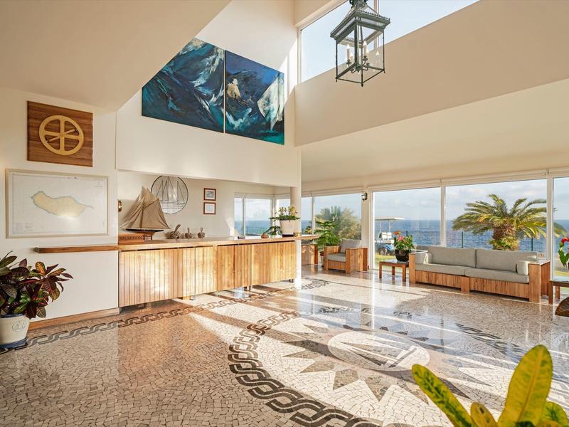 Jasna lobby hotelowa z recepcją i miejscem do siedzenia z widokiem na morze i roślinami.