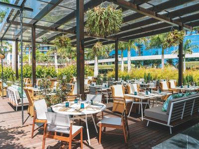 Světá terasa s stolky a židlemi pod skleněnou střechou, obklopená zelení.