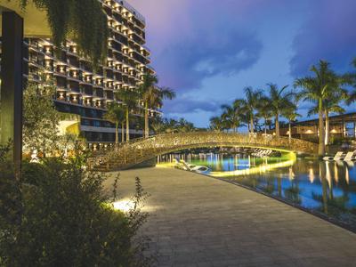 Oświetlona ścieżka hotelowa obok basenu z mostkiem i palmami o zmierzchu.