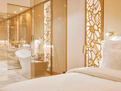 Světle hotelový pokoj s bílou postelí a moderní koupelnou se skleněnou stěnou.