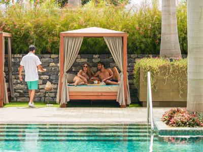 Dvě osoby odpočívají na zastřešeném lehátku u bazénu, třetí osoba přichází.