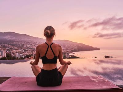 Žena meditujíc na jógové podložce s výhledem na klidnou vodu a město při západu slunce.
