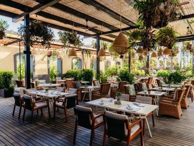 Krytá venkovní restaurace s dřevěnou podlahou, stoly, židlemi a mnoha závěsnými rostlinami.