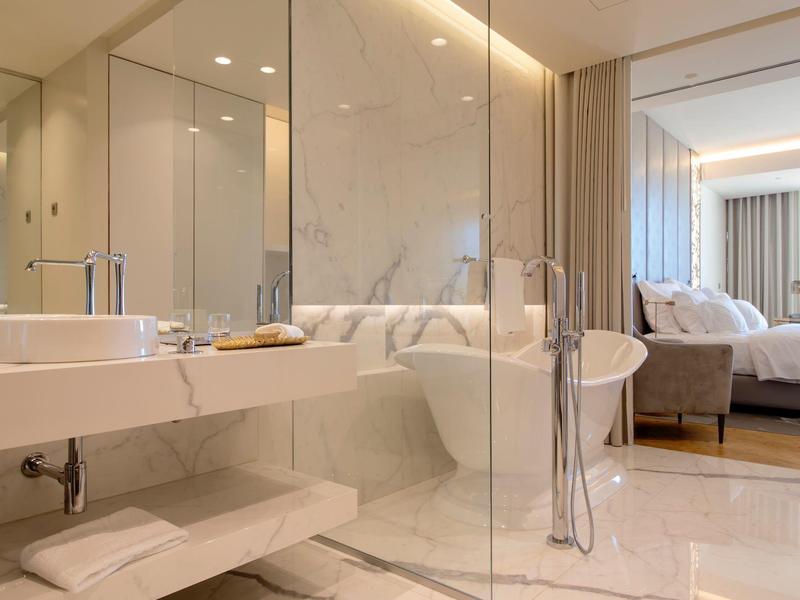 Moderní koupelna s vanou, skleněným sprchovým koutem a elegantním umyvadlem přiléhající k ložnici.