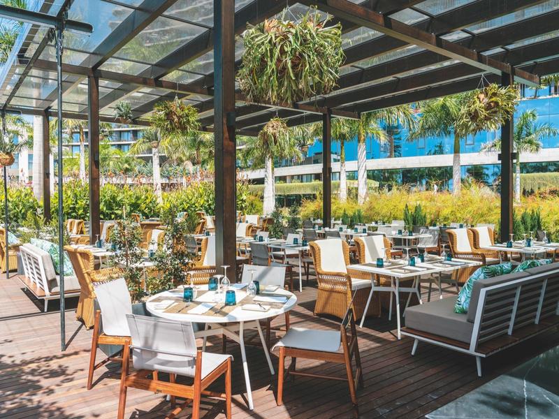 Světá terasa s stolky a židlemi pod skleněnou střechou, obklopená zelení.