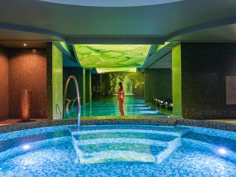 Vnitřní bazén s osvětlenými mozaikovými dlaždicemi a odpočívající osobou v pozadí