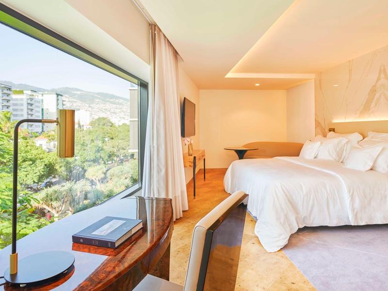 Světý hotelový pokoj s velkým oknem, útulnou postelí a výhledem na zelené stromy a budovy.