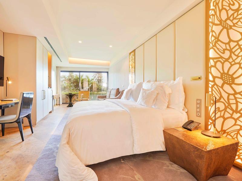 Světelný hotelový pokoj s velkou postelí, psacím stolem, televizí a dekorativní příčkou.