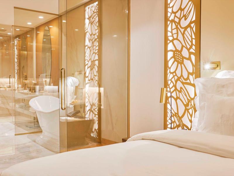 Světle hotelový pokoj s bílou postelí a moderní koupelnou se skleněnou stěnou.